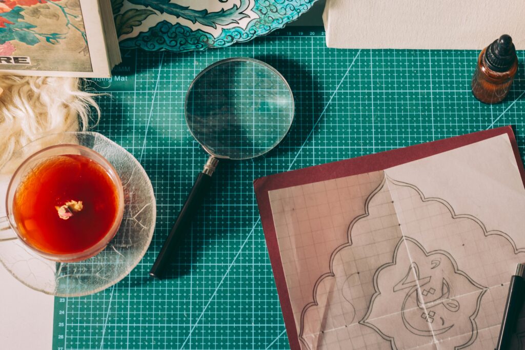 A cozy workspace with art supplies, tea, and a magnifying glass on a cutting mat.